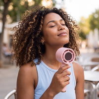 Light-skinned Black woman with natural curly hair cooling off at an outdoor cafe with the Frostbite mini turbo fan in soft pink
